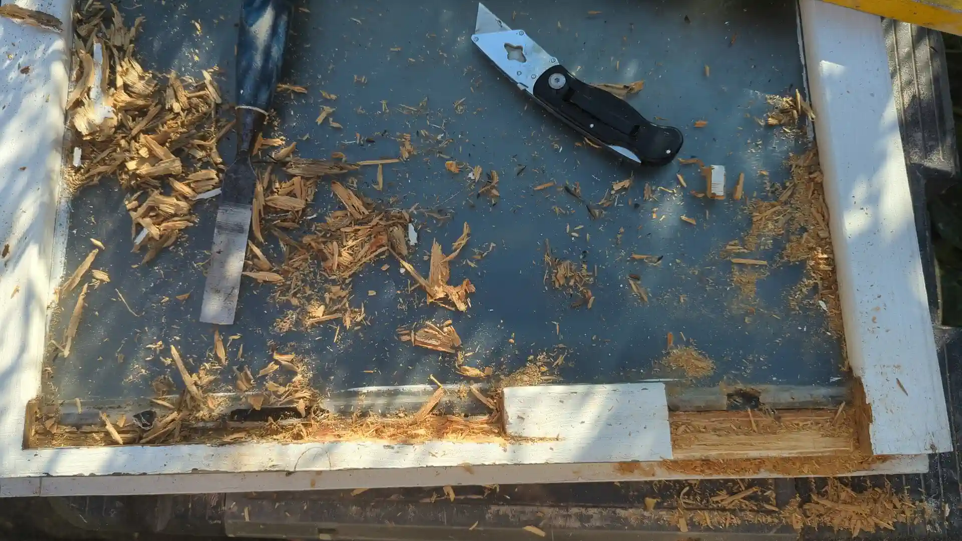Rotten timber chiselled out of a casement window bottom rail, with wood chips and a chisel on the glass